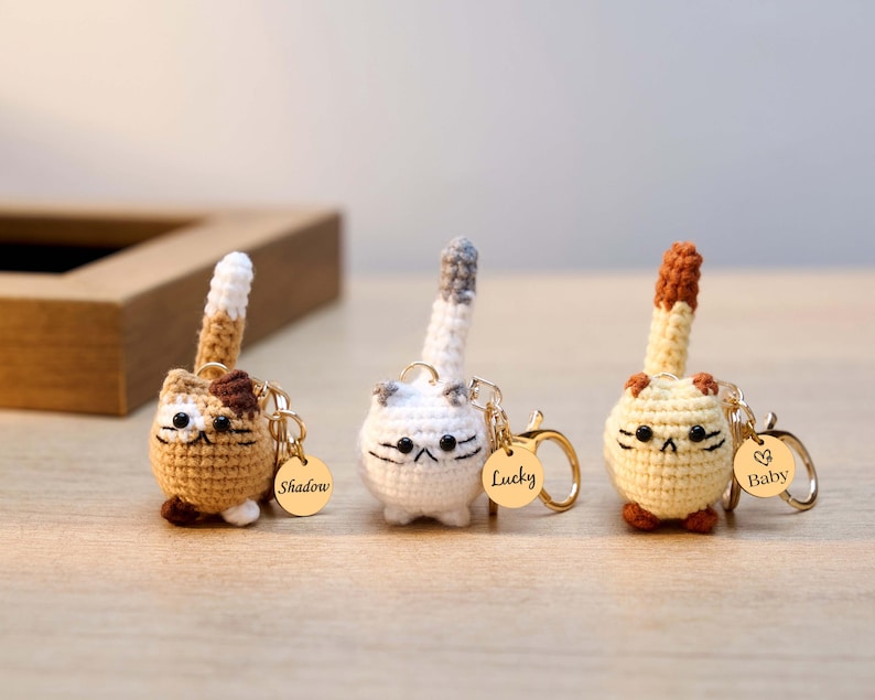 May include: Three handmade crochet cat keychains in brown, white, and yellow. Each cat has a gold-colored keychain ring and a small tag with a name. The cats have black embroidered facial features and upright tails.