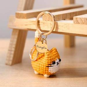 May include: A crocheted orange cat keychain with a gold-colored keyring and a circular tag that reads "Pumpkin." The cat has white accents and black eyes. The keychain is displayed in front of a wooden structure.