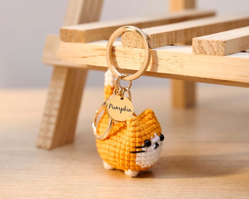 May include: A crocheted orange cat keychain with a gold-colored keyring and a circular tag that reads "Pumpkin." The cat has white accents and black eyes. The keychain is displayed in front of a wooden structure.