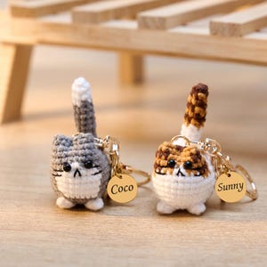 May include: Two crocheted cat keychains. One is gray and white with a gray and white tail, the other is brown, white, and tan with a brown and tan tail. Each keychain has a gold-colored tag with a name.