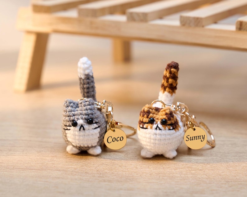 May include: Two crocheted cat keychains. One is gray and white with a gray and white tail, the other is brown, white, and tan with a brown and tan tail. Each keychain has a gold-colored tag with a name.