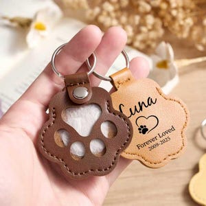 May include: Two leather paw-shaped keychains. One is brown with a paw print cutout, the other is tan with the name "Luna", a heart, paw print, and "Forever Loved 2009-2025" printed on it. Both have silver key rings.