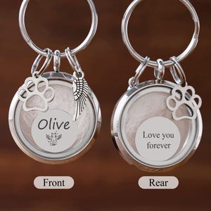 May include: Two silver-toned memorial keychains. Each keychain has a round, clear locket with a paw print charm. One keychain has the name "Olive" and a small angel graphic. The other says "Love you forever".