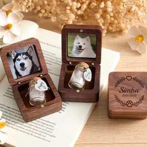 Peut inclure: Boîtes commémoratives en bois pour animaux de compagnie avec photos de chiens. Une boîte fermée, gravée "Simba 2013-2025". Deux boîtes ouvertes, chacune avec une photo et un petit flacon en verre. Un flacon porte une étiquette "Happy", l'autre "Lucky".