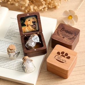 May include: Wooden pet memorial boxes with engraved names and paw prints. One box is open, displaying a photo of a golden retriever and a small glass vial with a heart-shaped tag that says "Happy". Other boxes are closed, with names and dates.