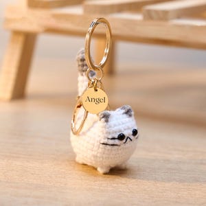 May include: A white crochet cat keychain with gray accents on the ears and tail. The keychain has a gold-colored ring and a small, round tag that reads "Angel". The cat has black eyes and a black stitched mouth.