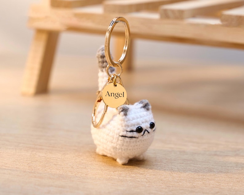 May include: A white crochet cat keychain with gray accents on the ears and tail. The keychain has a gold-colored ring and a small, round tag that reads "Angel". The cat has black eyes and a black stitched mouth.
