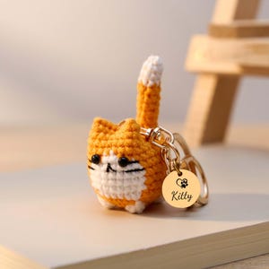 May include: A crocheted orange and white cat keychain with a gold-colored tag. The tag has a heart and paw print design with the name "Kitty" engraved. The cat has black eyes and a white muzzle.