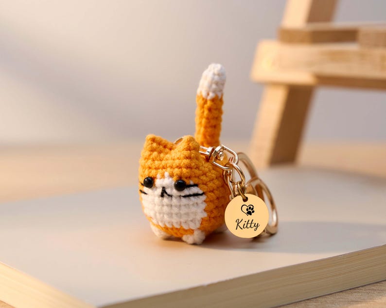 May include: A crocheted orange and white cat keychain with a gold-colored tag. The tag has a heart and paw print design with the name "Kitty" engraved. The cat has black eyes and a white muzzle.
