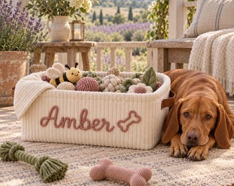 May include: A rectangular cream-colored corduroy storage basket with brown leather handles. The front features the name Amber and a bone icon in raised pink yarn. The basket is filled with various crocheted toys, including a bee and flowers.