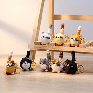 May include: A collection of handmade crochet cat keychains in various colors, including white, black, gray, orange, and brown. Each keychain features a small gold tag with a name. The keychains are displayed on a wooden shelf.