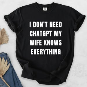 Può includere: T-shirt nero con la scritta bianca "I DON'T NEED CHATGPT MY WIFE KNOWS EVERYTHING". La maglietta è a girocollo e sembra essere realizzata in un materiale morbido. La maglietta è piegata, con un paio di jeans blu e una pianta secca decorativa sullo sfondo.