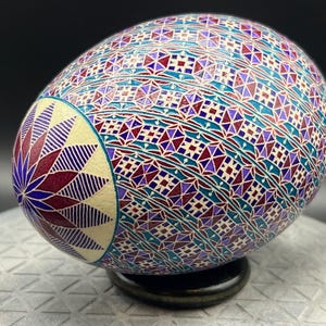 May include: A decorative egg with intricate geometric patterns in teal, purple, burgundy, and cream. The egg is displayed on a black stand, showcasing the detailed artwork. The design features a floral motif on one end.