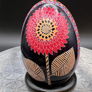 May include: A decorative, hand-painted egg with a large red and yellow sunflower design on a black background. The egg sits on a black stand. The sunflower has detailed petals and a patterned centre, with tan leaves and stem.