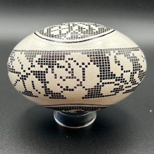 Op de afbeelding: Een ovaalvormig decoratief object met een crèmekleurige basis, versierd met een zwart geometrisch patroon. Het ontwerp heeft een mozaïekachtige opstelling van kleine vierkantjes en lijnen. Het object wordt op een transparante standaard gepresenteerd.