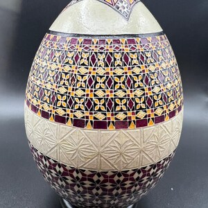 May include: An egg-shaped decorative object with a cream-colored top and base. The body features a mosaic pattern of burgundy, yellow, and black geometric shapes. The base has a carved starburst design.