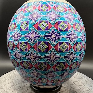 Op de afbeelding: Een decoratief, bolvormig object met een gedetailleerd, geometrisch patroon. Het ontwerp bevat een combinatie van turquoise, paarse, roze en groene vormen, waaronder ruiten en sterren. Het bovenste gedeelte is crèmekleurig en het object staat op een zwarte basis.