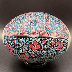 Könnte beinhalten: Aufwendig verzierte Eierschale mit floralem und geometrischem Design. Der obere Bereich zeigt ein Muster aus roten, blauen und schwarzen geometrischen Formen. Der untere Bereich zeigt ein Blumendesign mit roten Blüten und blauen Blättern auf schwarzem Hintergrund.