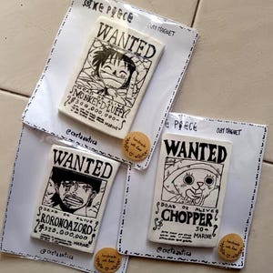 Può includere: Magneti in argilla fatti a mano con personaggi di One Piece. Ogni magnete presenta un design di poster "RICERCATO" con illustrazioni di personaggi e taglie. I magneti sono su cartoncini bianchi con bordi decorativi.