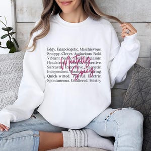 May include: White sweatshirt with the words "Tenacity" in pink script. The sweatshirt has a list of words in black, including "Edgy," "Mischievous," and "Passionate."