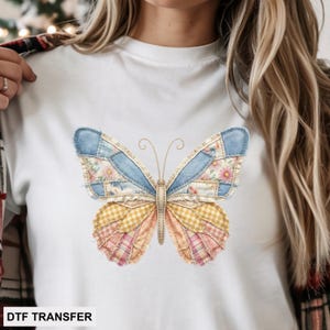 May include: White t-shirt featuring a colorful butterfly design. The butterfly has patchwork wings in shades of blue, pink, yellow, and floral patterns. The text "DTF TRANSFER" is printed at the bottom of the shirt.