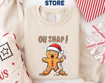 May include: A cream-colored sweatshirt with a graphic of a gingerbread man wearing a Santa hat and holding a candy cane. The text "OH SNAP!" is above the gingerbread man. The sweatshirt is surrounded by Christmas-themed decorations.