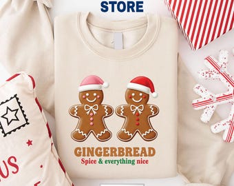 May include: Beige sweatshirt featuring two gingerbread men wearing Santa hats, with the text "GINGERBREAD Spice & everything nice." The design is festive and holiday-themed, perfect for Christmas.