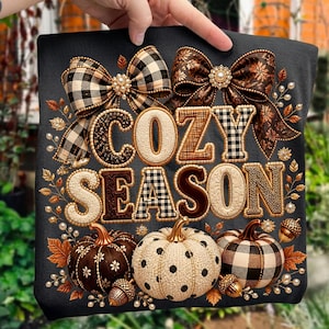 Op de afbeelding: Een zwart t-shirt met de tekst COZY SEASON in gestructureerde, decoratieve letters. Het ontwerp bevat twee grote strikken, herfstbladeren, parels en drie pompoenen met bruine, witte en zwarte ruitpatronen en stippen.