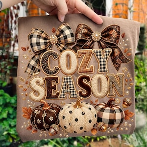 Op de afbeelding: Een beige t-shirt met de tekst COZY SEASON in gestructureerde, gedessineerde letters. Het ontwerp bevat twee grote strikken aan de bovenkant, drie pompoenen aan de onderkant en verspreide herfstbladeren, eikels en parels.