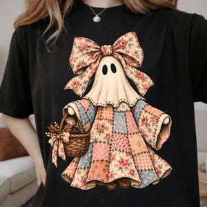 May include: A black t-shirt featuring a graphic of a ghost wearing a large floral bow and a patchwork dress made of various pink, blue, and orange patterned squares. The ghost holds a woven basket filled with autumn leaves and a matching bow.