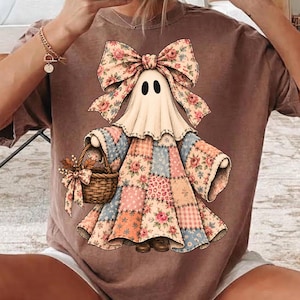 Può includere: Una t-shirt marrone con la grafica di un fantasma che indossa un grande fiocco floreale e un abito patchwork con vari motivi, tra cui rose e quadretti. Il fantasma tiene in mano un piccolo cesto intrecciato decorato con un fiocco floreale.