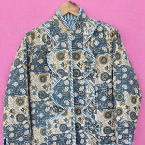 Peut inclure: Veste matelassée à motifs floraux dans les tons de bleu, beige et vert olive. La veste a un col montant et est bordée d'un passepoil bleu clair. Les manches ont un poignet boutonné. La veste est suspendue à un cintre en bois.