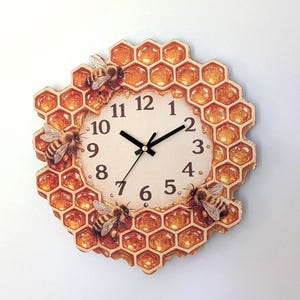 May include: A round wall clock with a cream-coloured face featuring black Arabic numerals and black hands. The frame is shaped like a honeycomb structure with amber-coloured resin cells and three detailed honeybee figures attached to the border.