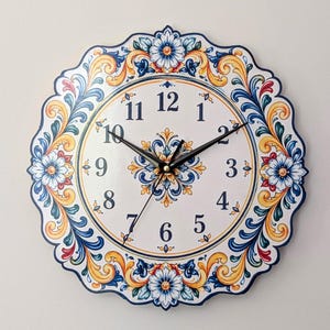 May include: A round wall clock with a scalloped edge featuring a white face and black numerals from 1 to 12. The border and centre are decorated with intricate floral and scroll patterns in shades of blue, yellow, and red on a white background.