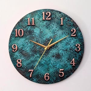 May include: A round wall clock with a textured, mottled teal and black surface. The clock features raised copper-coloured numerals from one to twelve and three thin, gold-coloured hands indicating the hour, minute, and second positions.