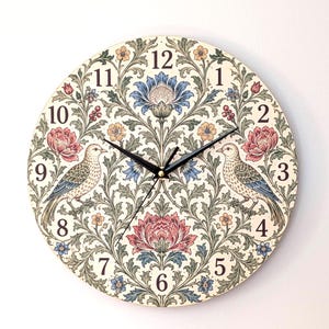 May include: A round wall clock featuring a cream-coloured background with a symmetrical botanical pattern of green leaves, blue and pink flowers, and two birds facing each other. The clock has black numerals from 1 to 12 and thin black clock hands.