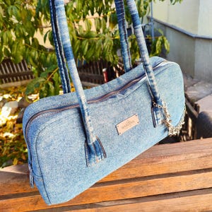 Peut inclure: Sac à main en jean bleu clair de forme rectangulaire avec fermeture éclair. Le sac est doté de deux anses et d'une petite étiquette métallique portant l'inscription "MARIANA". Une chaîne argentée avec des breloques en forme d'étoiles est suspendue sur le côté.