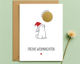 May include: A Christmas card featuring a white rabbit in a Santa hat gazing upwards at a gold circle. The card includes the words "FROHE WEIHNACHTEN" and a small red heart. It is presented inside a brown envelope.