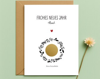 May include: A white greeting card with the text "FROHES NEUES JAHR" and a name written below. The card features a gold circle surrounded by a black and white floral wreath, a red heart, and the text "Kreis freirubbeln". The card is in a brown envelope.