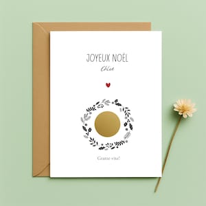 May include: A white Christmas card with the words "JOYEUX NOËL" and "Chloe" in black script. A red heart is above a gold scratch-off circle surrounded by a black and white wreath. The card is in a brown envelope.