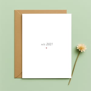 May include: A white card with the text "seit 2021" and a small red heart, inside a brown envelope. A single flower with a beige bloom and a long stem is placed on the right side of the card. The background is a light green colour.