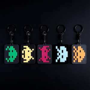 Op de afbeelding: Vijf sleutelhangers met gepixelde alien-ontwerpen in groen, geel, rood, lichtblauw en oranje. Elke sleutelhanger heeft een zwarte achtergrond en een zwarte metalen ring. De ontwerpen doen denken aan retro videogames.