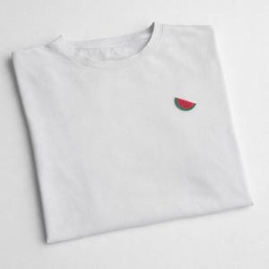 May include: White t-shirt with a small embroidered watermelon slice on the chest. The watermelon design features red and green colors. The shirt is made of a soft, lightweight material, perfect for casual wear.