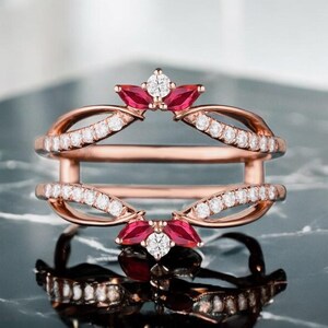 May include: Rose gold ring enhancer with a double band design. The ring features small white diamonds and red gemstones in a floral arrangement at the top and bottom. The ring is displayed on a reflective surface.