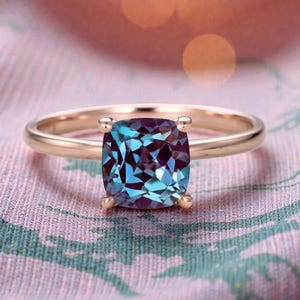 Può includere: Un anello d'oro con una grande pietra preziosa a taglio quadrato che cambia colore. La pietra preziosa mostra sfumature di blu e viola. L'anello è posto su uno sfondo di tessuto fantasia.