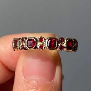 May include: A gold ring featuring a series of vibrant red gemstones. The ring's design incorporates a mix of square and round-cut stones, creating a unique and eye-catching pattern. The gemstones are set in a gold band.