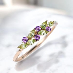 Puede incluir: Un delicado anillo de oro con un diseño floral. El anillo está adornado con piedras preciosas de peridoto de corte ovalado en un tono verde claro y piedras preciosas de amatista de corte redondo en color púrpura. Las piedras están engarzadas en una fila a lo largo de la banda del anillo.