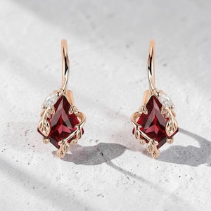 May include: A pair of rose gold earrings featuring a square, deep red gemstone. Each earring has a leaf design and a small, clear accent stone. The earrings are displayed against a light background.