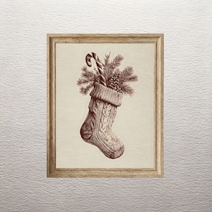 May include: A sepia-toned framed print of a Christmas stocking filled with pine needles, a candy cane, and a pine cone. The stocking has a cable knit design. The frame is made of wood.