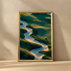 May include: A framed print depicting a golf course from above. The image shows a meandering sand trap, verdant green fairways, and a golf cart. The frame is gold-coloured, and the overall feel is tranquil and natural.